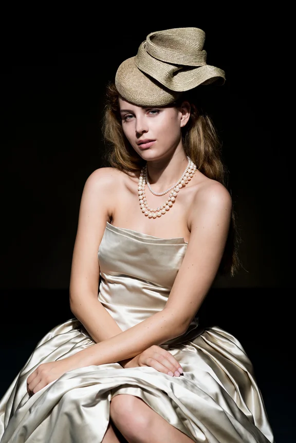 sitzende weibliche Person in silbernem schulterfreiem Kleid mit Hut und Halskette